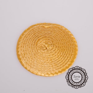 May include: A circular, golden-yellow woven straw hat base with a flat, spiraled design. The material features a consistent braided texture throughout the entire surface. A black circular logo with the text Zoria is visible in the corner.