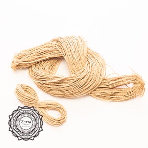 May include: A bundle of light tan, fibrous twine arranged in a loose, coiled loop with a smaller separate coil nearby. A circular black and white logo featuring the text Zoria and decorative filigree patterns is positioned in the lower left corner.