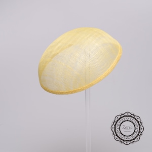 May include: A pale yellow oval fascinator hat featuring a woven mesh texture and a structured brim. The hat has a smooth, rounded dome shape and is displayed on a clear stand. A circular logo with the text Zoria is visible in the corner.