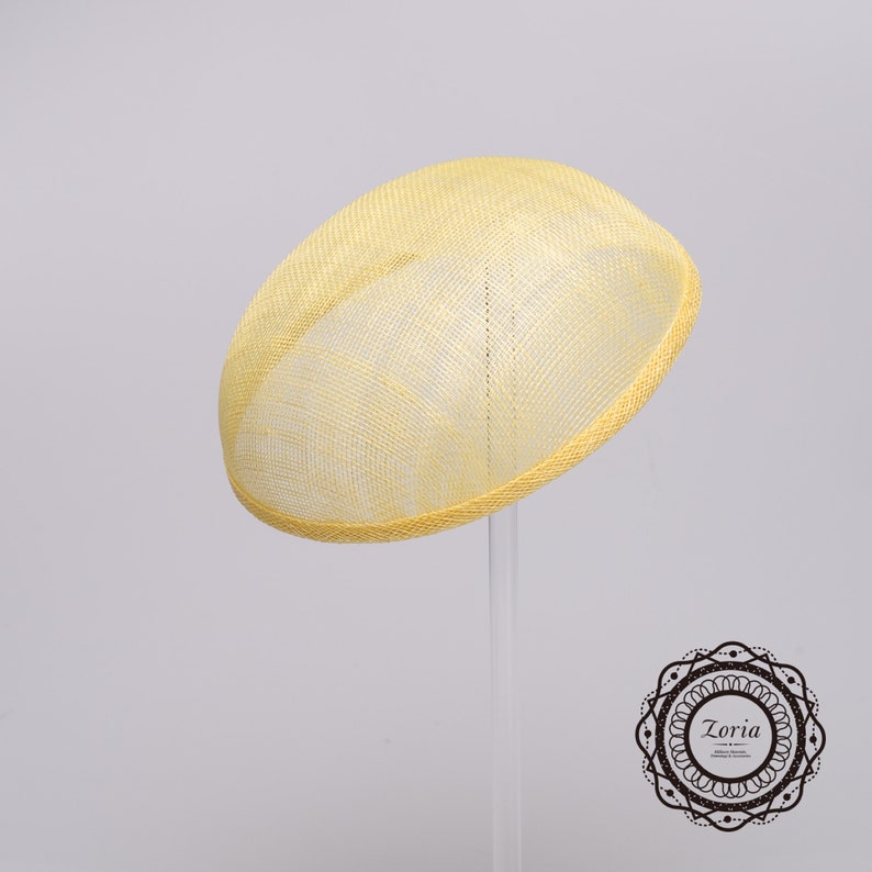 May include: A pale yellow oval fascinator hat featuring a woven mesh texture and a structured brim. The hat has a smooth, rounded dome shape and is displayed on a clear stand. A circular logo with the text Zoria is visible in the corner.