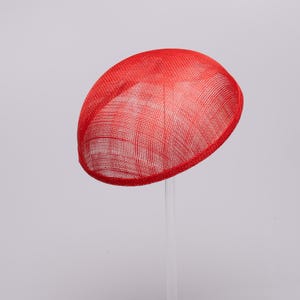 May include: A bright red fascinator hat with a rounded, dome-shaped crown and a stiff, woven mesh texture. The hat is mounted on a clear, vertical support stand, showcasing its smooth, curved silhouette and vibrant, solid red color.