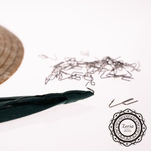 May include: A close-up of a pile of silver-colored metal hooks, a dark green fabric strip with a hook at the end, and a woven straw hat. The hooks are scattered on a white surface.