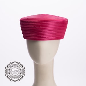 May include: A vibrant fuchsia pillbox hat featuring a structured, cylindrical shape with horizontal ribbed detailing throughout the exterior. The hat is displayed on a neutral mannequin head, showcasing its solid, bright pink color and clean lines.