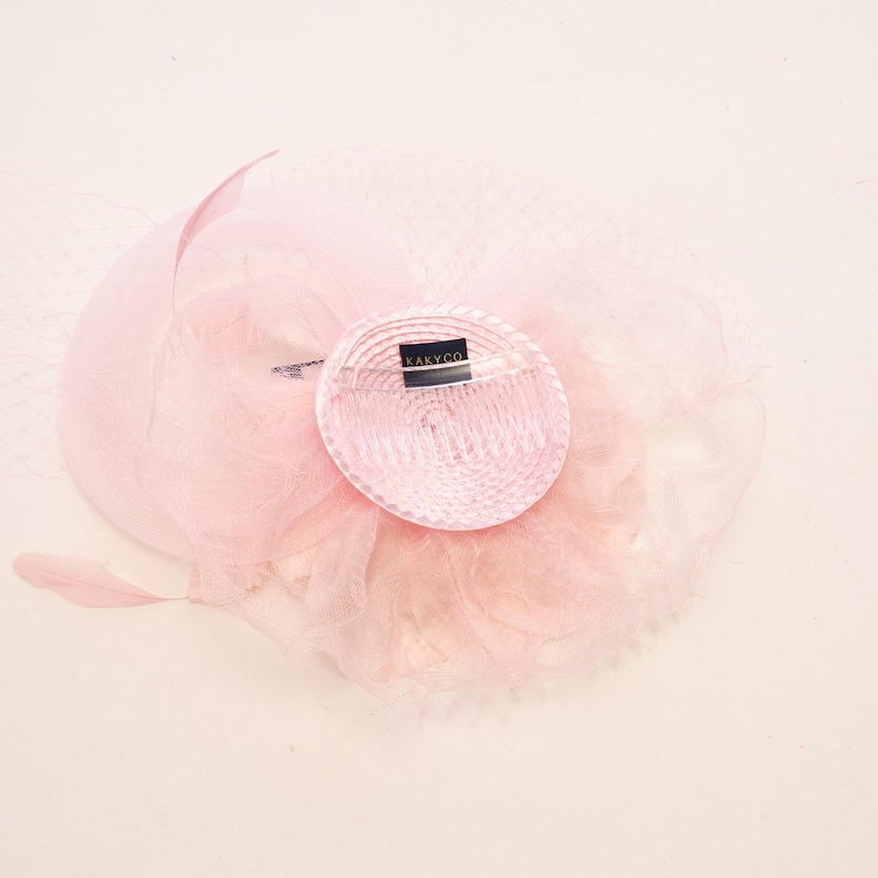 May include: A light pink fascinator hat featuring a circular base with a ribbed texture, a black label with the text KAKYCO, layered tulle netting, and a single feather accent. The accessory is constructed with delicate, sheer fabric elements.