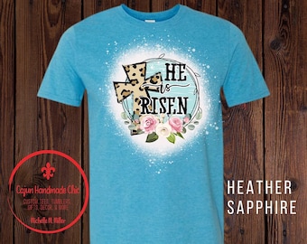 May include: A light blue t-shirt with a white and blue floral design and a leopard print cross. The text "HE is RISEN" is written in a white circle with a blue background. The shirt is made of a soft, comfortable material.