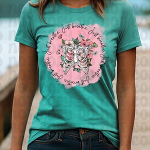 Pu&ograve; includere: Maglietta turchese con un grafico circolare rosa che presenta una gabbia toracica stilizzata con accenti floreali. Il testo "Just breathe. Just breathe. Just breathe" &egrave; scritto attorno al cerchio.