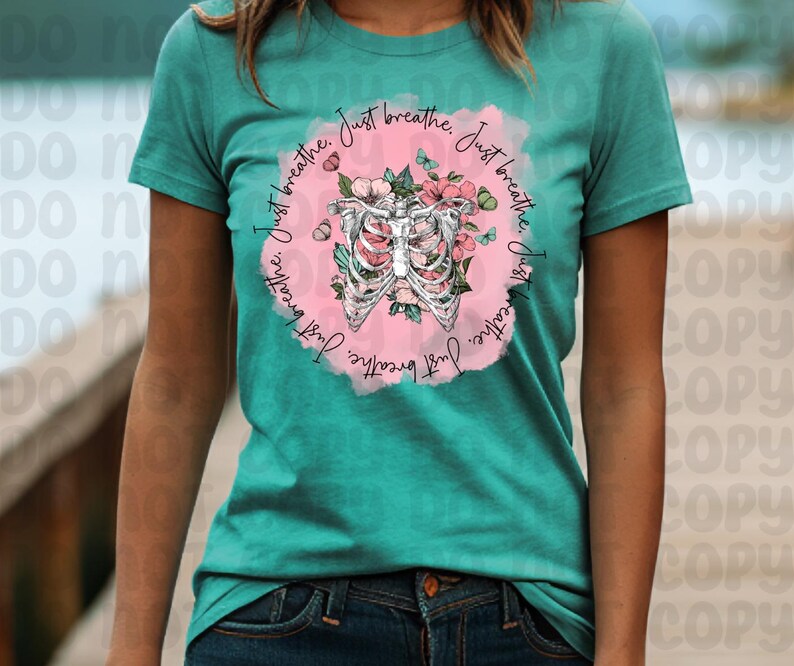 Pu&ograve; includere: Maglietta turchese con un grafico circolare rosa che presenta una gabbia toracica stilizzata con accenti floreali. Il testo "Just breathe. Just breathe. Just breathe" &egrave; scritto attorno al cerchio.