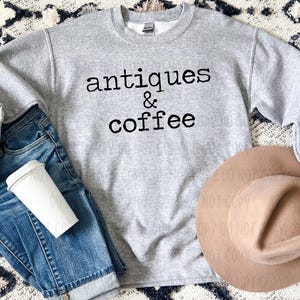 May include: A heather gray sweatshirt with the words "antiques & coffee" in black lettering. The sweatshirt is paired with blue jeans, a white coffee cup, and a tan hat. The image has a casual, lifestyle theme.