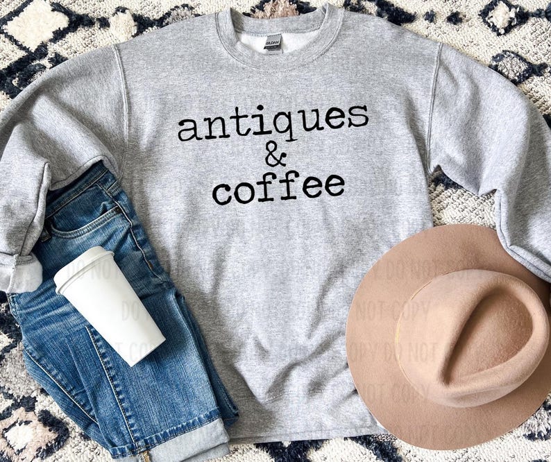 May include: A heather gray sweatshirt with the words "antiques & coffee" in black lettering. The sweatshirt is paired with blue jeans, a white coffee cup, and a tan hat. The image has a casual, lifestyle theme.