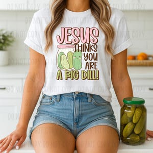 May include: White t-shirt with the text "JESUS THINKS YOU ARE A BIG DILL!" in pink and brown. The graphic features a jar of pickles with a smiling pickle. The shirt is paired with denim shorts.