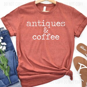 May include: A rust-colored t-shirt with the words "antiques & coffee" in white, displayed with blue jeans, white flowers, and brown sandals. The shirt is folded at the bottom.