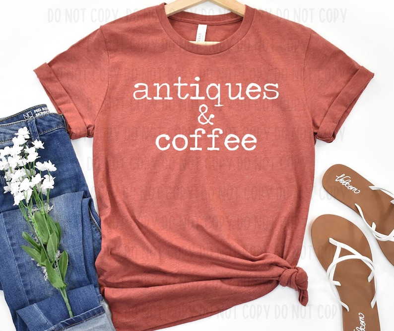 May include: A rust-colored t-shirt with the words "antiques & coffee" in white, displayed with blue jeans, white flowers, and brown sandals. The shirt is folded at the bottom.