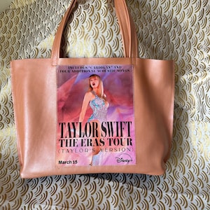 May include: A light brown leather tote bag with a clear vinyl patch featuring a concert poster for Taylor Swift's The Eras Tour. The poster includes a photo of Taylor Swift, the tour dates, and the Disney+ logo. The text on the poster reads "INCLUDES 'CARDIGAN' AND FOUR ADDITIONAL ACOUSTIC SONGS TAYLOR SWIFT THE ERAS TOUR (TAYLOR'S VERSION) March 15 Disney+"
