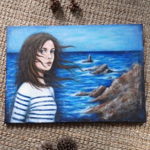 Puede incluir: Pintura al óleo de una mujer con cabello castaño largo y ojos azules, vestida con una camisa a rayas, mirando al espectador. El fondo presenta un paisaje marino con un faro, rocas y un cielo azul.
