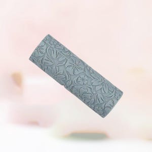 May include: A gray, cylindrical rolling pin with a floral pattern. The surface is covered in embossed flowers and leaves, creating a textured design. The rolling pin is designed for baking and crafting.