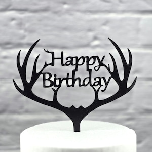May include: Black cake topper with the words "Happy Birthday" and a deer antler design. The topper is placed on top of a white cake, with a gray brick wall in the background.