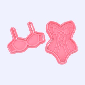 May include: Two pink plastic cookie cutters in the shape of lingerie. One is a bra, and the other is a corset. The bra has straps and cups. The corset has a detailed design with lacing and boning.
