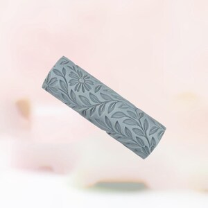 May include: A gray, cylindrical rolling pin with a floral and leaf pattern. The design features a central daisy surrounded by detailed leaves and vines. The rolling pin is made of a solid material and is designed for baking.