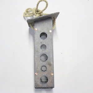 May include: A rectangular grey bird feeder with a flat roof and a thick beige rope for hanging. The front panel features five circular openings covered with a fine metal mesh grid, secured by four small copper-coloured rivets at the corners.