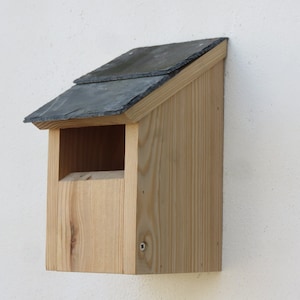 May include: A wooden birdhouse with a slate roof. The birdhouse is light brown and has a small opening for birds to enter.