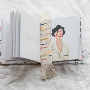 Op de afbeelding: Een wit boek met een bloemenmotief op de kaft is open op een pagina met de tekst "In de late schemering zijn de schaduwen lang en diep". Een kleurrijk geweven bladwijzer met een franje hangt aan het boek.