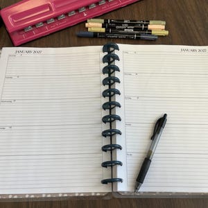 May include: An open disc-bound planner with white lined pages dated January 2027. A black retractable pen rests on the right page. A pink hole punch and three dual-tip markers in blue, light yellow, and light green are positioned above the planner.