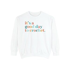 May include: White long-sleeve sweatshirt with the text "it's a good day to crochet." in colorful letters.