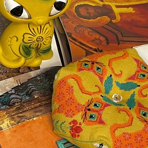 May include: A yellow and orange needlepoint pillow with a design of four cats. The pillow is sitting on a book with the text "Villa Country" visible.