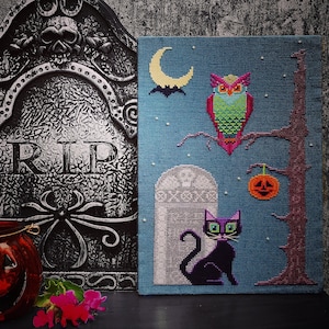 May include: A cross-stitch embroidery design featuring a black cat, an owl, and a pumpkin. The design is on a blue background with a tombstone and a tree. The text "RIP" is on a black tombstone.
