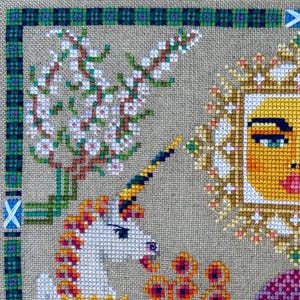 Könnte beinhalten: Kreuzstich-Stickerei mit einem weißen Einhorn mit Regenbogenmähne und -horn, einer blühenden Blume und einer Sonne mit einem menschlichen Gesicht. Der Hintergrund ist ein beigefarbener Stoff mit einem grün-blauen karierten Rand.