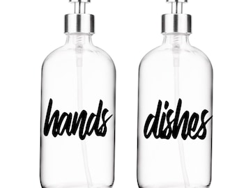 May include: Two clear glass soap dispensers with silver pumps. The dispenser on the left has a black label that says "hands" and the dispenser on the right has a black label that says "dishes".