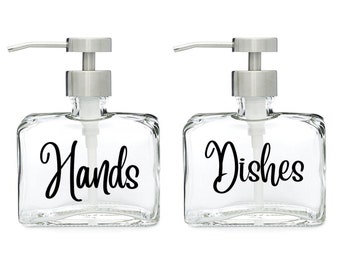 May include: Two clear glass soap dispensers with stainless steel pumps. The dispenser on the left is labeled "Hands" and the dispenser on the right is labeled "Dishes".