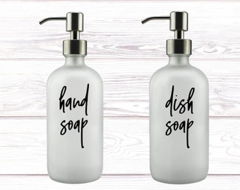 May include: Two frosted glass soap dispensers with silver pumps. The dispenser on the left says "hand soap" and the dispenser on the right says "dish soap".