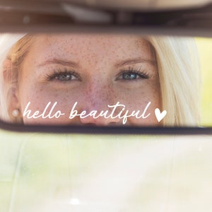 Peut inclure: Le reflet d'un visage de femme dans un rétroviseur. La réflexion montre une femme souriante avec les yeux ouverts. Le texte "hello beautiful" avec un symbole de cœur est écrit sur le miroir.