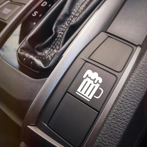 May include: A black car interior detail featuring a button with a white beer mug icon. The button is part of a control panel near the gear shift. The image is a close-up, highlighting the design and placement of the button.