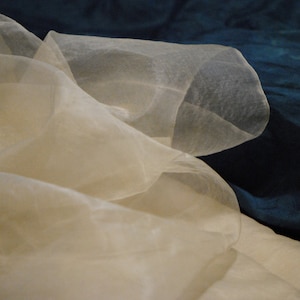 May include: A sheer, white fabric with a delicate, flowing texture. The fabric is draped over a dark blue surface.