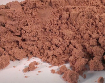 May include: Close-up of a pile of fine, reddish-brown powder. The powder is textured and appears to be a bulk product, with a clear plastic bag visible in the background. The powder is spread out on a white surface.