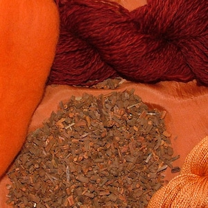 May include: A close-up of a pile of dried, brown, natural dye material.  There are two skeins of yarn, one orange and one dark red, and a piece of orange fabric in the background.
