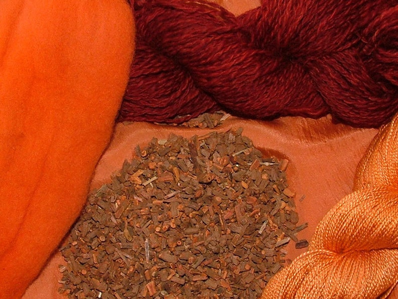 May include: A close-up of a pile of dried, brown, natural dye material.  There are two skeins of yarn, one orange and one dark red, and a piece of orange fabric in the background.