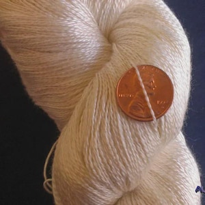 May include: Close-up of a skein of off-white yarn. A U.S. penny is placed on top of the yarn for scale. The yarn appears to be soft and smooth, with a slight sheen. The yarn is likely used for knitting or crocheting.