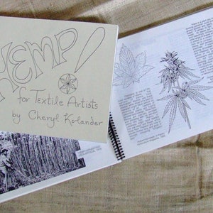 May include: A closed book titled "Hemp! for Textile Artists by Cheryl Kolander" with a spiral binding. The cover features a stylized drawing of the word "Hemp!" and a small illustration of a hemp leaf. The book is open to a page with a line drawing of a hemp plant.