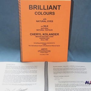 May include: An orange spiral-bound book titled "BRILLIANT COLOURS" with the subtitle "with NATURAL DYES". The book is open to a page with text and illustrations. The author's name, Cheryl Kolander, is visible, along with the Aurora Silk logo.