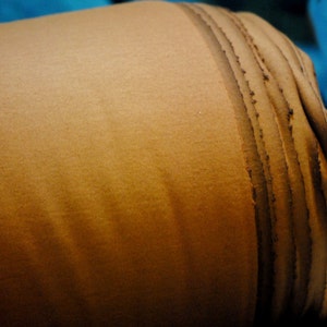 May include: A close-up of a brown fabric roll with a textured surface. The fabric is rolled up tightly and has a dark brown seam running along the edge.