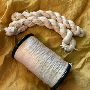 May include: A spool of natural colored thread with a black plastic bobbin, and a loose bundle of the same thread on a yellow fabric background.