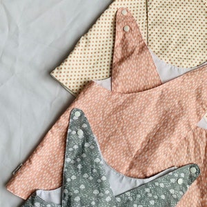 May include: Three fabric baby bibs with different patterns. The top bib is white with brown polka dots, the middle bib is pink with a white mushroom pattern, and the bottom bib is gray with a white mushroom pattern.