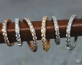 May include: A set of six dainty rings with a single row of clear cubic zirconia stones. The rings are arranged in a row on a wooden surface, with the metal colors ranging from silver to rose gold.