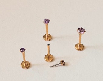 May include: Five gold body jewelry pieces with purple gemstones. Four pieces are studs with a post and a base. One piece is a post only.