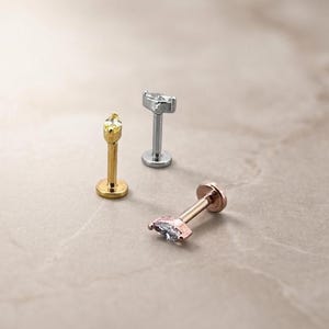 May include: Three flat-back labret studs featuring marquise-cut clear stones. One stud is gold-toned, one is silver-toned, and one is rose gold-toned. Each piece consists of a straight post, a circular base, and a decorative top with a single stone.