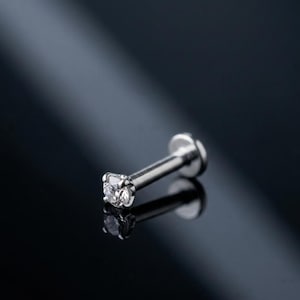 May include: A silver-toned labret body jewellery piece featuring a single clear, round-cut gemstone held in a four-prong setting at the end of a straight post with a flat circular disc base, reflecting light against a dark, smooth surface.