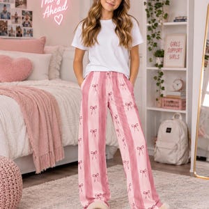 May include: A plain white short-sleeved t-shirt paired with loose-fitting pink and white striped pajama pants featuring a repeating pattern of small pink bows. The pants have an elastic drawstring waistband and side pockets, worn with tan slippers.